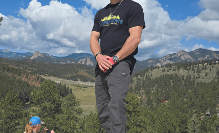 Person standing outdoors on a wooden surface with mountains and blue sky in background.
