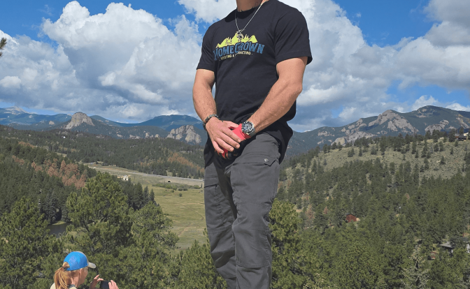 Person standing outdoors on a wooden surface with mountains and blue sky in background.
