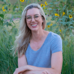 Woman with glasses and long blonde hair smiling outdoors among sunflowers and greenery.