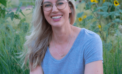 Woman with glasses and long blonde hair smiling outdoors among sunflowers and greenery.