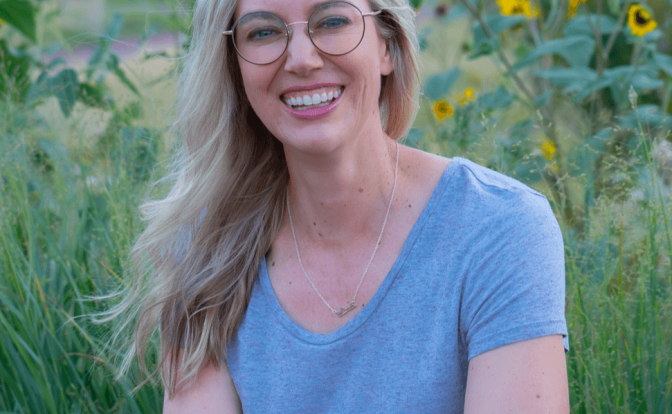 Woman with glasses and long blonde hair smiling outdoors among sunflowers and greenery.