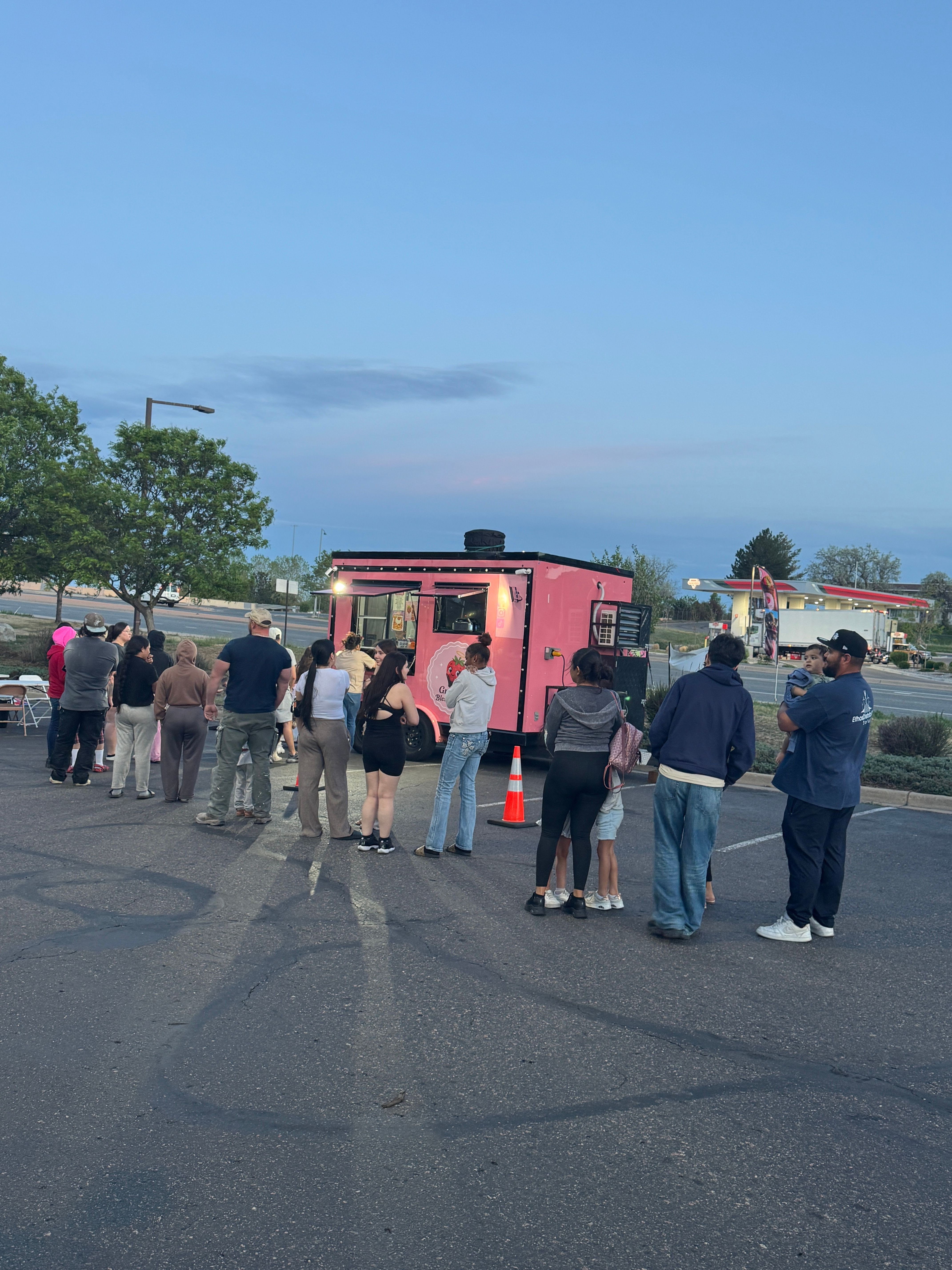 Group of people standing outdoors near a pink bus on a paved area, with trees and a blue sky in the background.