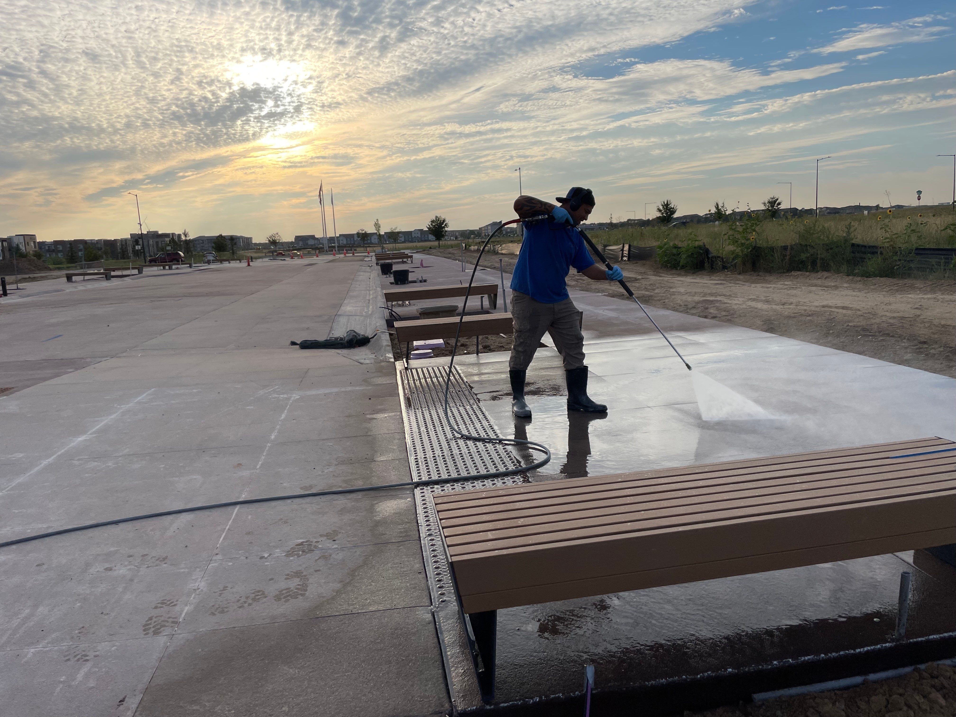 Person cleaning a large outdoor concrete surface with a hose, benches nearby, sky with clouds and sunlight, open area.
