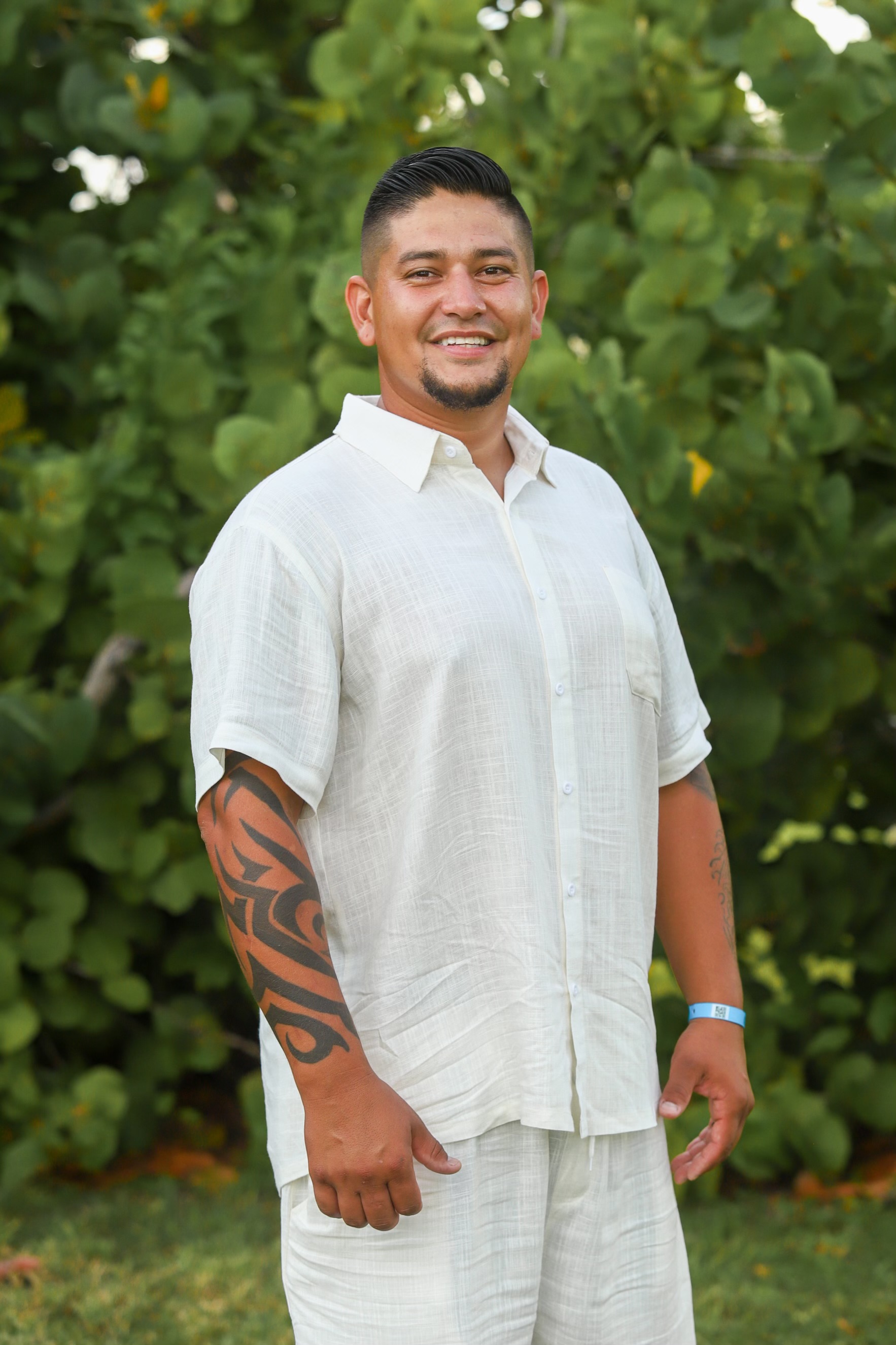 Man with short dark hair and a goatee, smiling, wearing a light-colored short-sleeve shirt and pants, standing outdoors with green foliage in background.