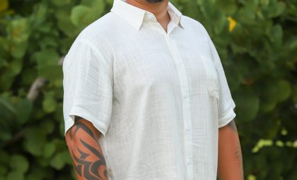 Man with short dark hair and a goatee, smiling, wearing a light-colored short-sleeve shirt and pants, standing outdoors with green foliage in background.