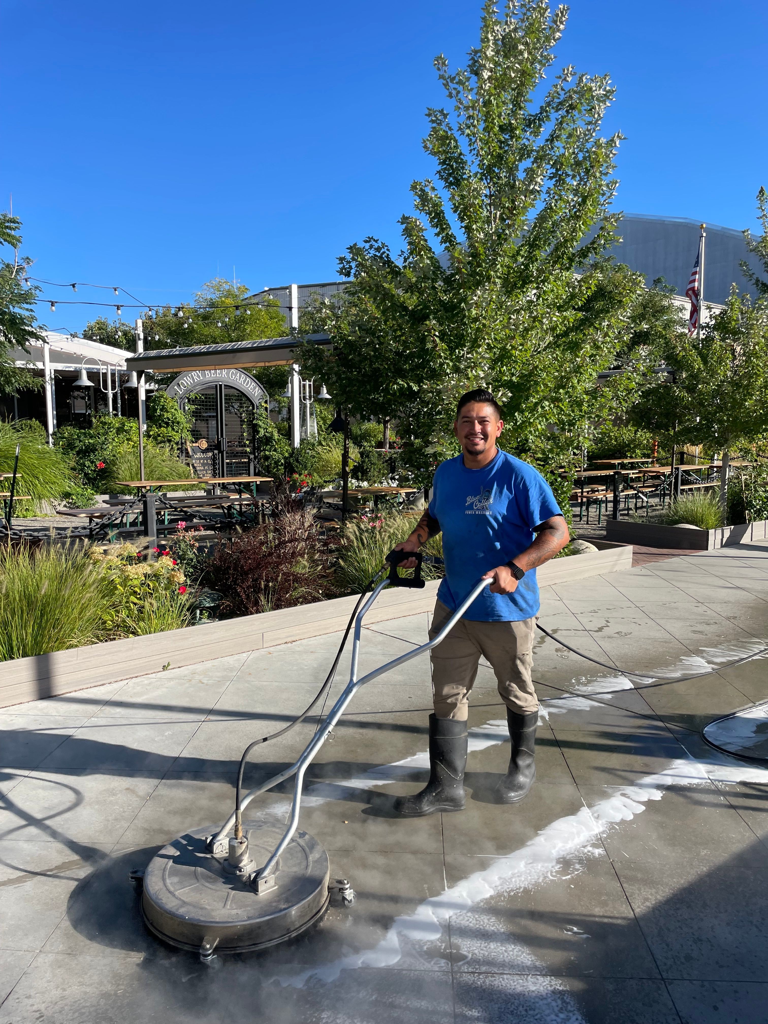 Man cleaning sidewalk with a power washer, trees and buildings in background, sunny day.