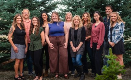 Group of ten women standing outdoors in front of trees, smiling, with a small green plant in the foreground.