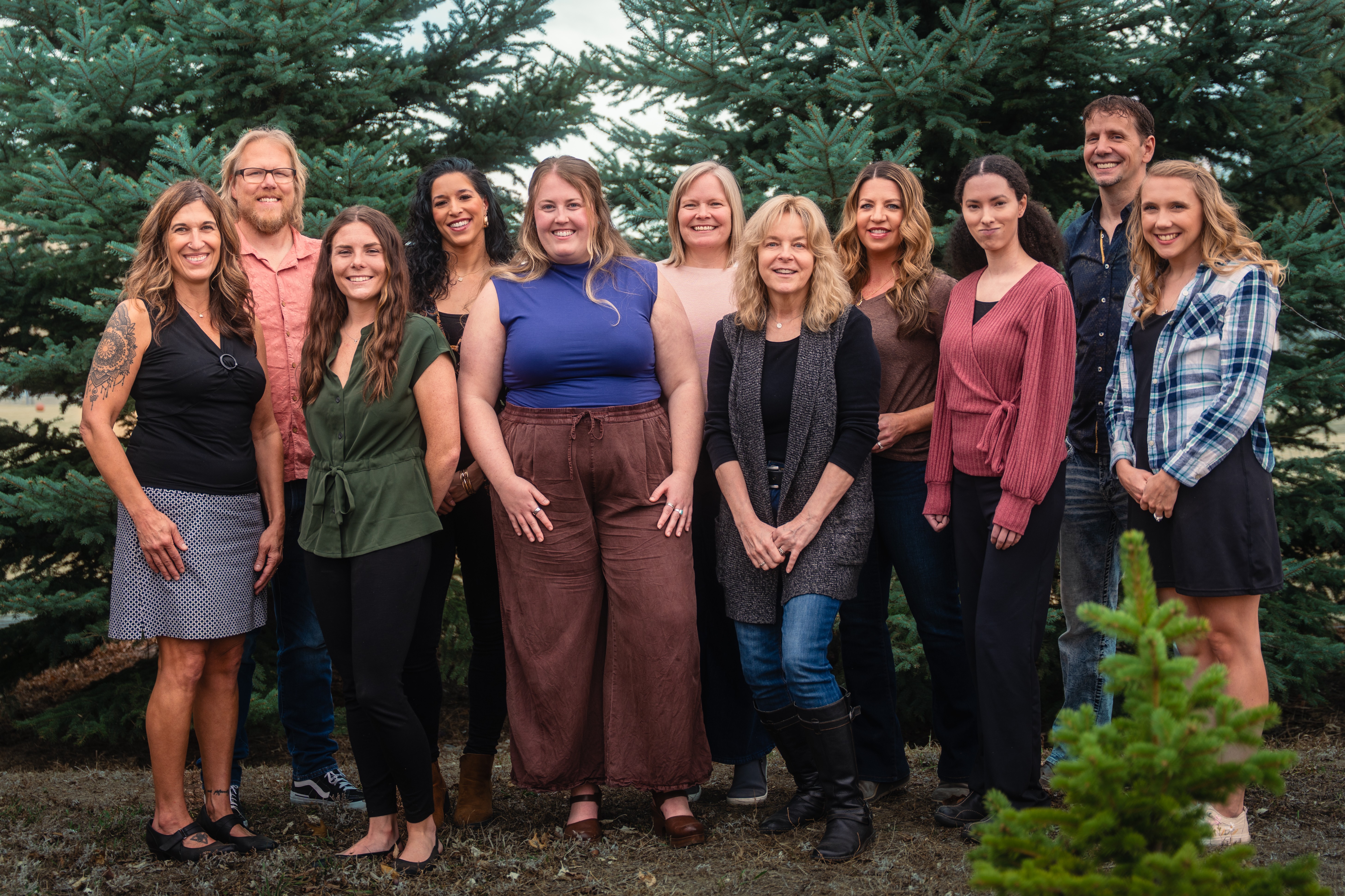 Group of ten women standing outdoors in front of trees, smiling, with a small green plant in the foreground.