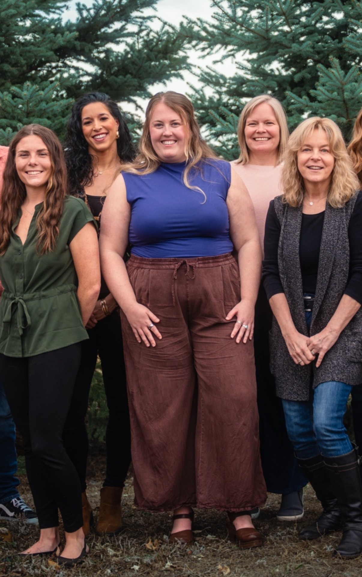 Group of five women standing outdoors among trees, smiling, dressed casually, with a natural background.