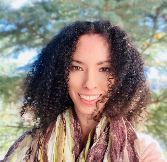 Smiling woman with curly hair outdoors, wearing a colorful scarf, trees in background.