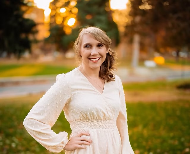 Woman with blonde hair smiling outdoors in a park during sunset, wearing a white dress with long sleeves.