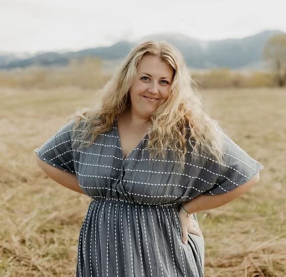 Woman with long curly blonde hair smiling outdoors in a field, wearing a blue and white striped dress.