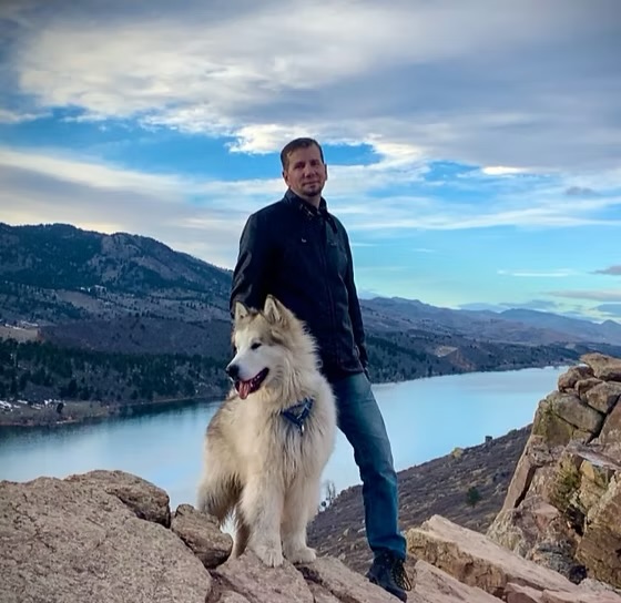 Man standing on rocks with a large fluffy dog near a river, mountains in the background, cloudy sky.
