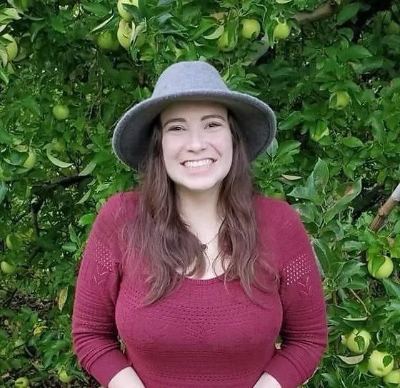 Young woman smiling, wearing a gray hat and maroon sweater, standing in front of green apple tree.