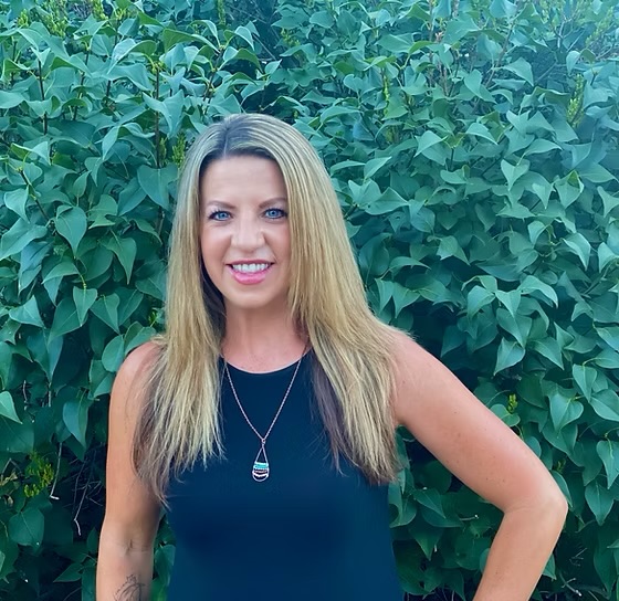 Woman with long blonde hair smiling outdoors in front of green leafy background.
