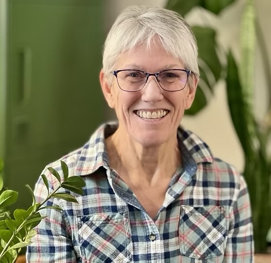 Smiling woman with short gray hair and glasses, wearing a plaid shirt, in a room with green plants.