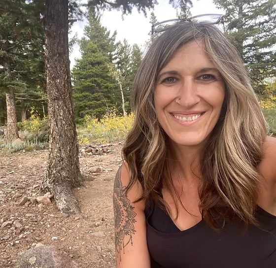 Smiling woman with wavy hair outdoors in a forested area, wearing a black top, with trees and yellow flowers in background.