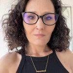 Woman with curly hair, glasses, and a nose piercing, wearing a black sleeveless top and a gold necklace.