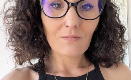 Woman with curly hair, glasses, and a nose piercing, wearing a black sleeveless top and a gold necklace.