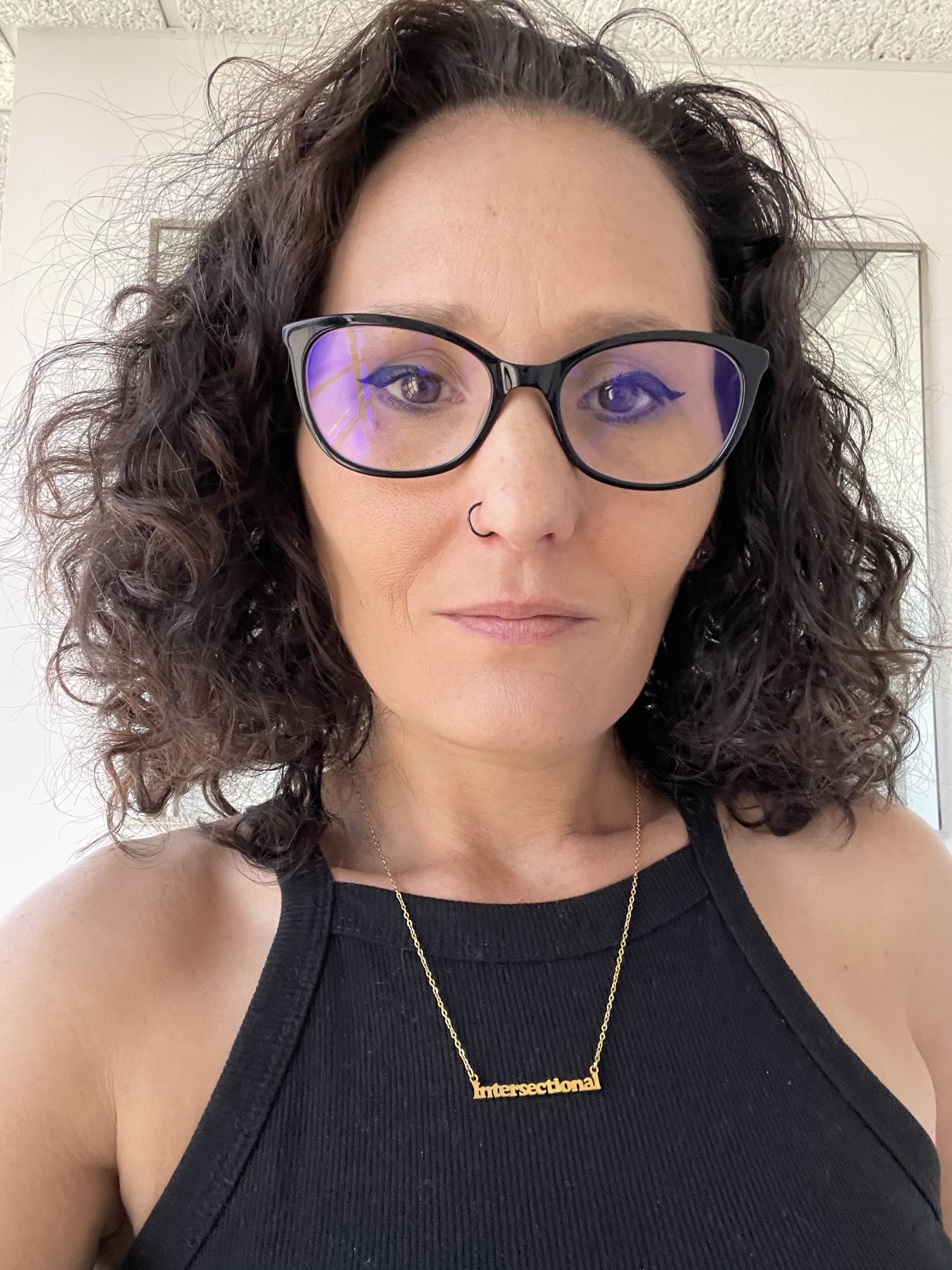 Woman with curly hair, glasses, and a nose piercing, wearing a black sleeveless top and a gold necklace.