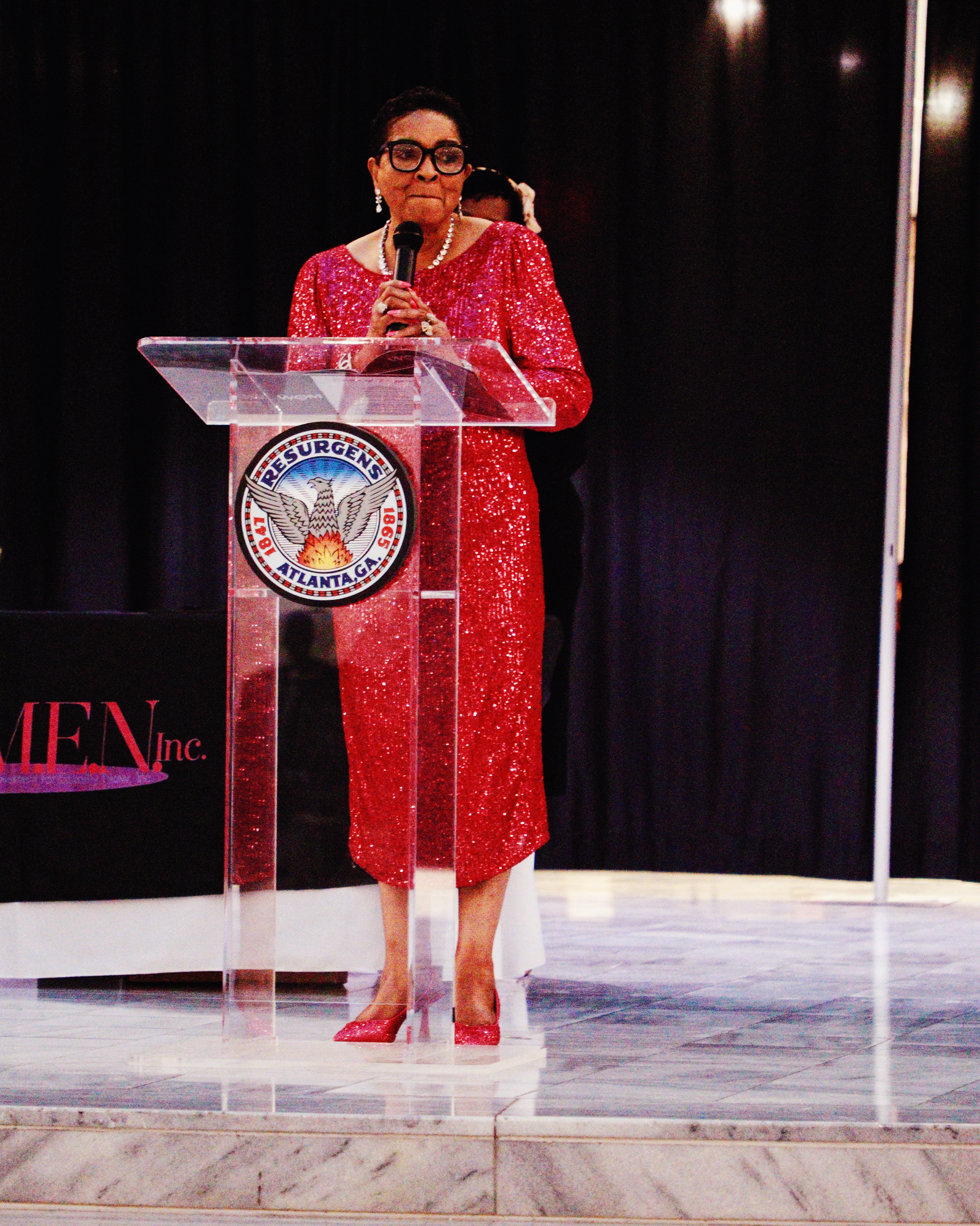 Woman in red dress speaking at a podium with a seal, black curtain background, holding a microphone.