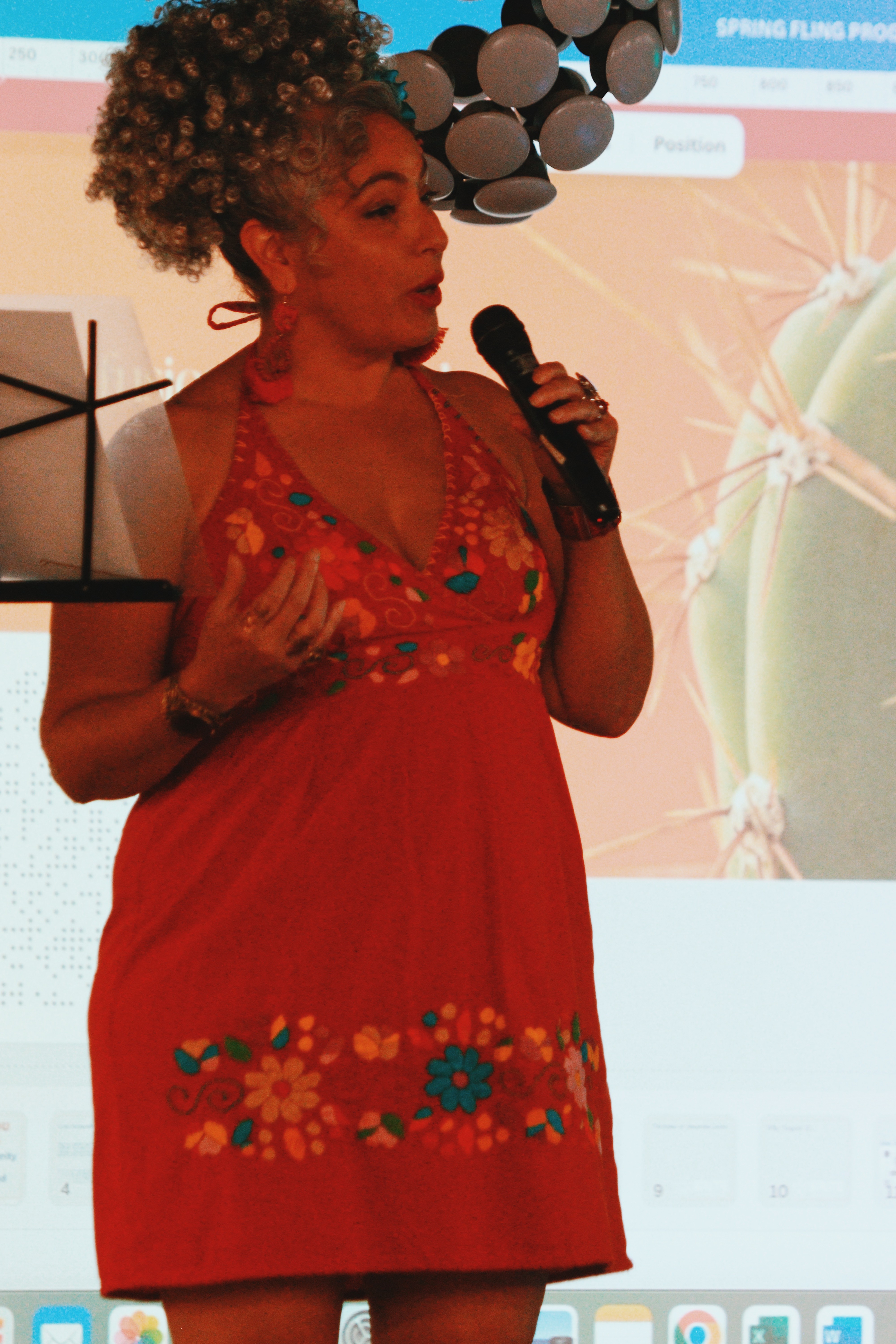 Woman with curly hair speaking into a microphone, wearing a red dress with floral embroidery, standing in front of a presentation screen.