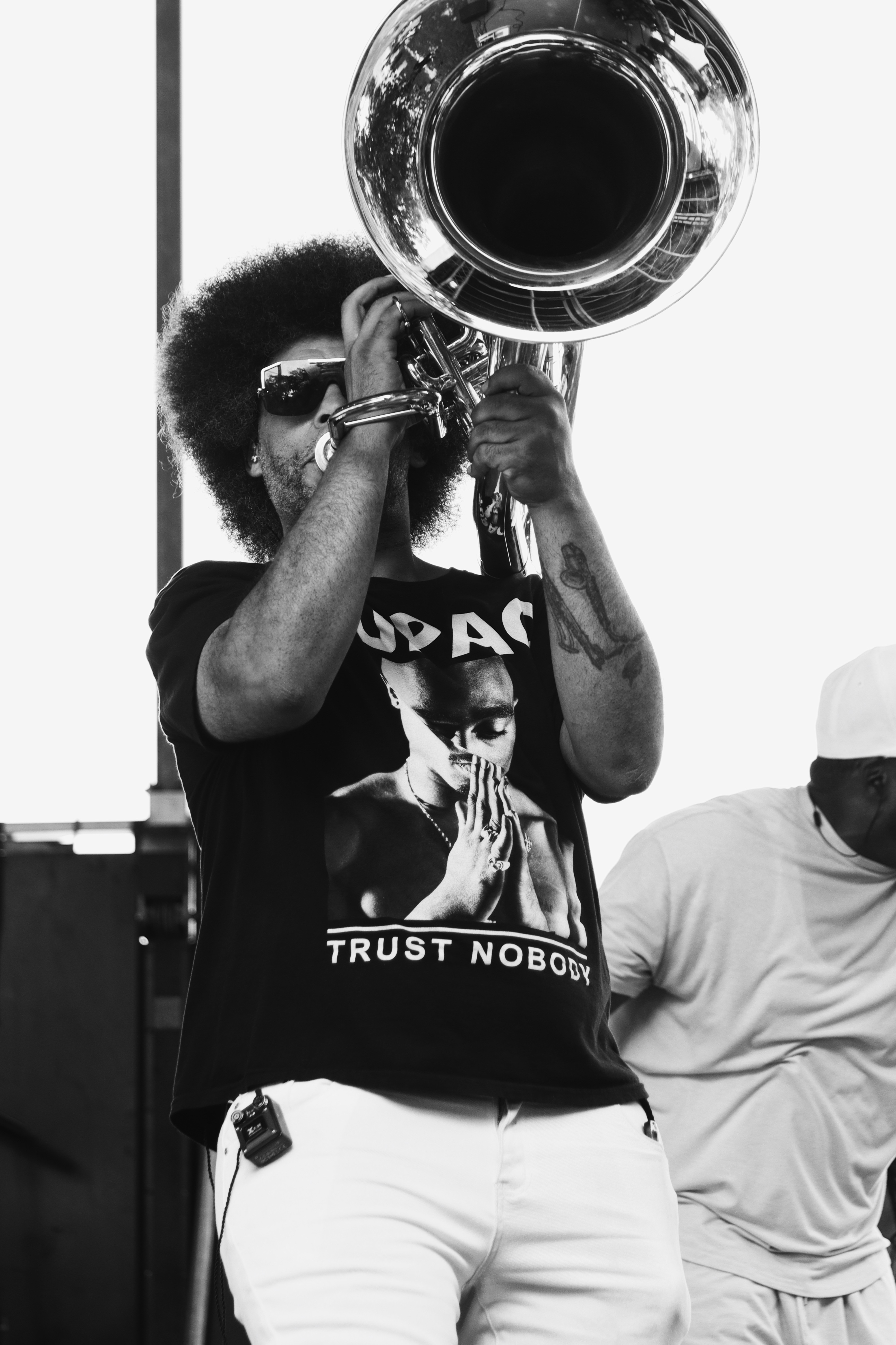 Person playing a large brass instrument, wearing sunglasses and a graphic T-shirt, with another person nearby, in black and white.