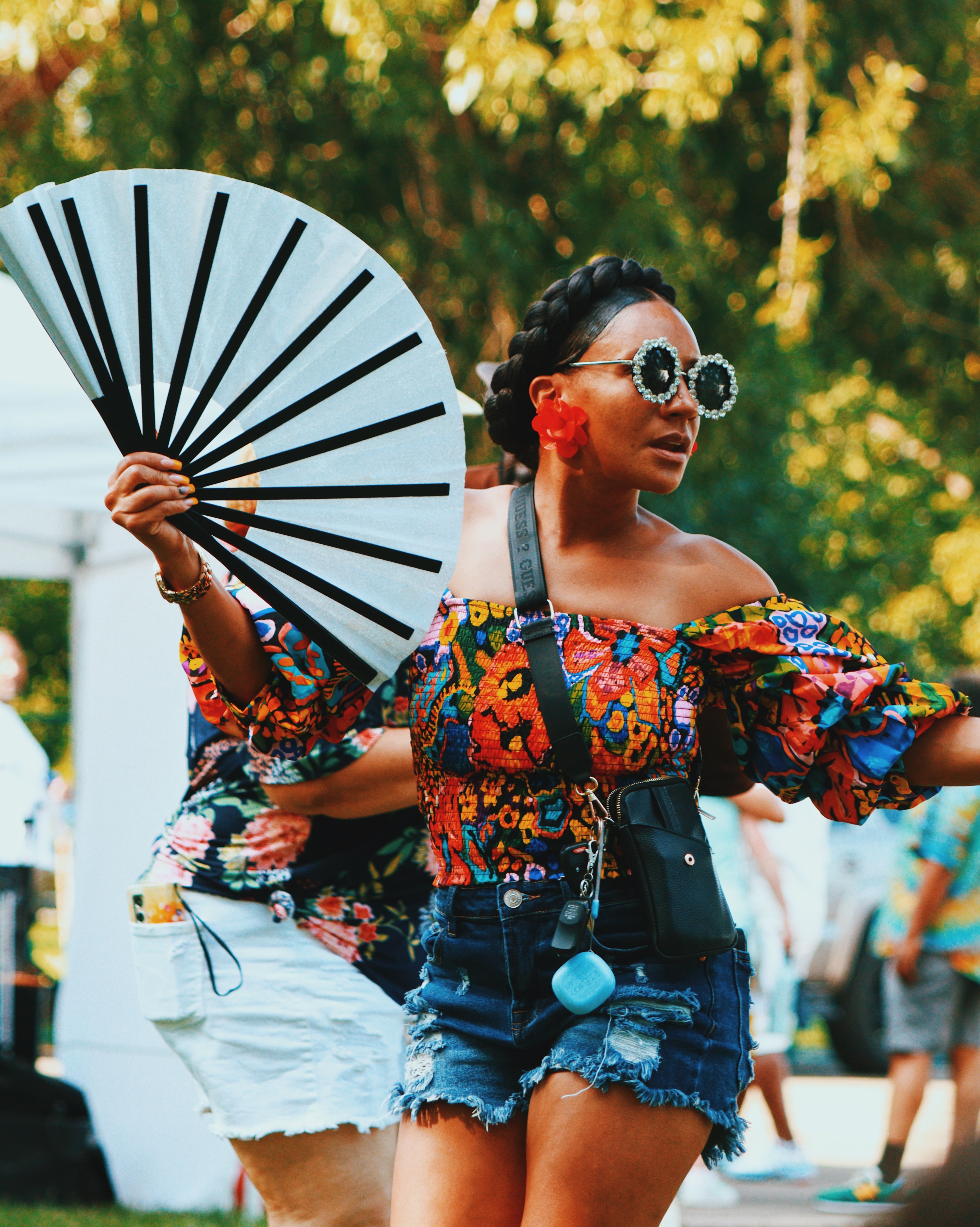 Woman wearing sunglasses and a colorful off-shoulder top, holding a large fan outdoors with trees in background.