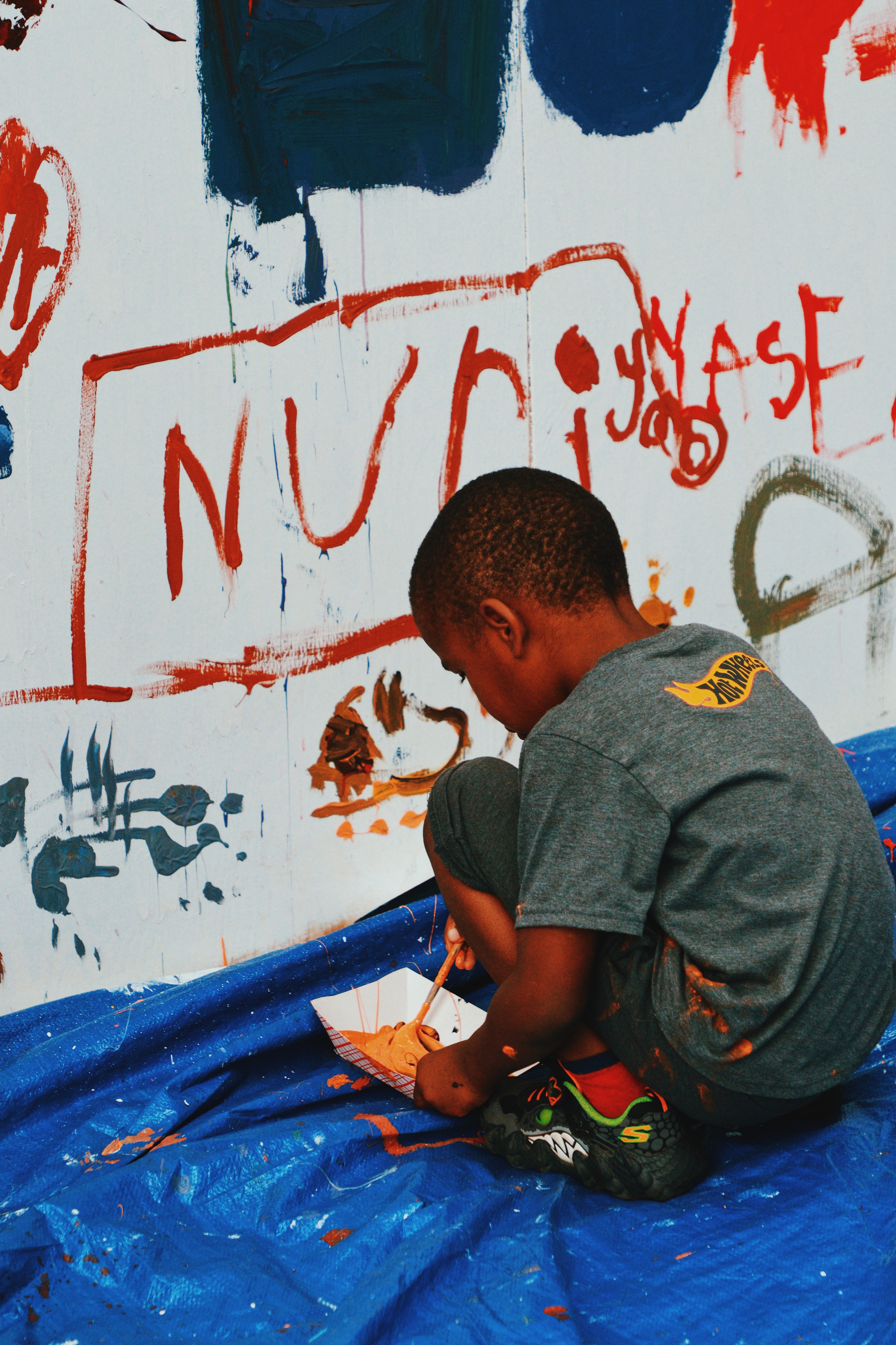 Child drawing on a wall with colorful paint and graffiti, sitting on a blue tarp.