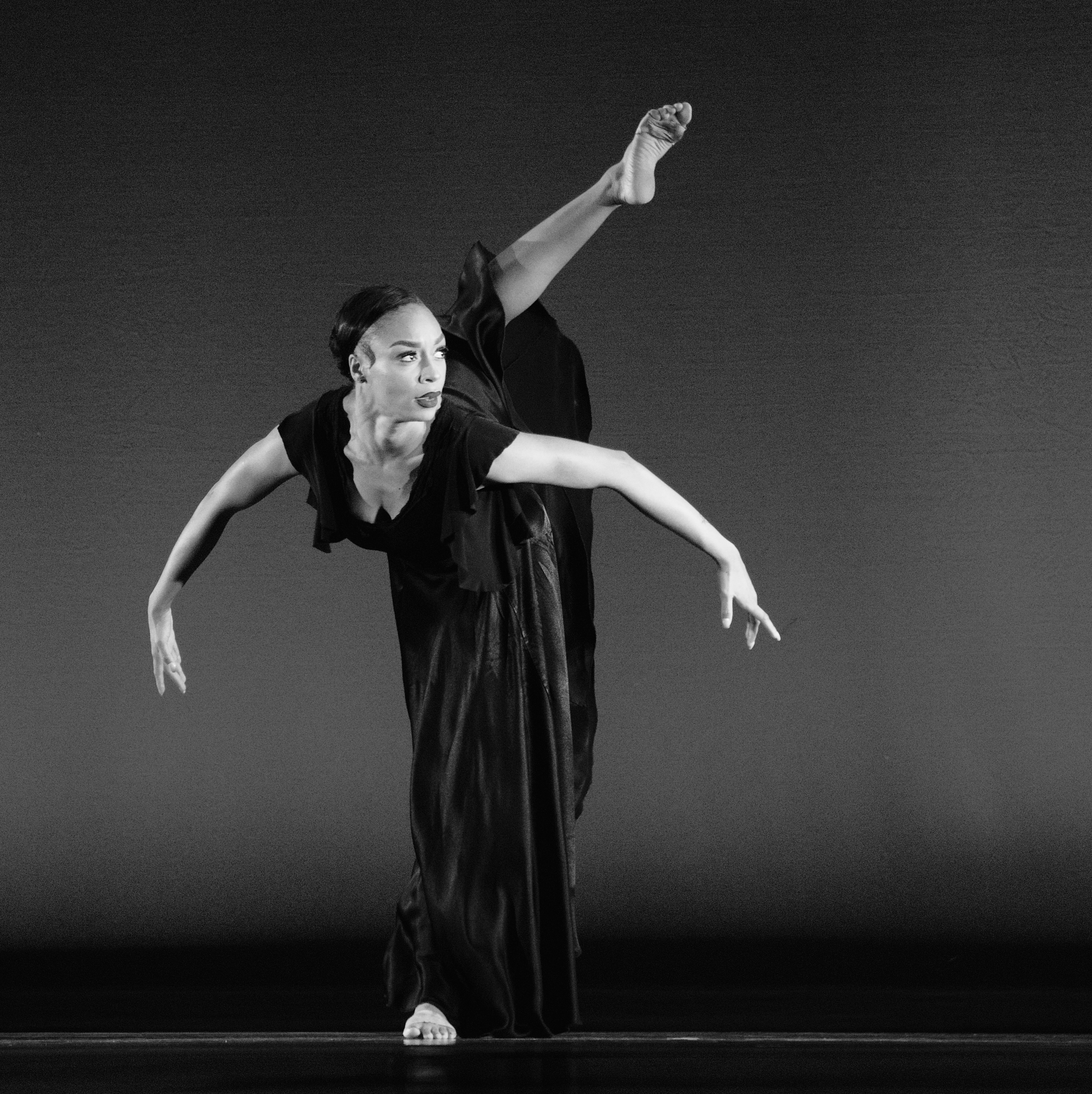 A dancer performs a pose with one leg raised and arms extended, on a stage with a plain background.