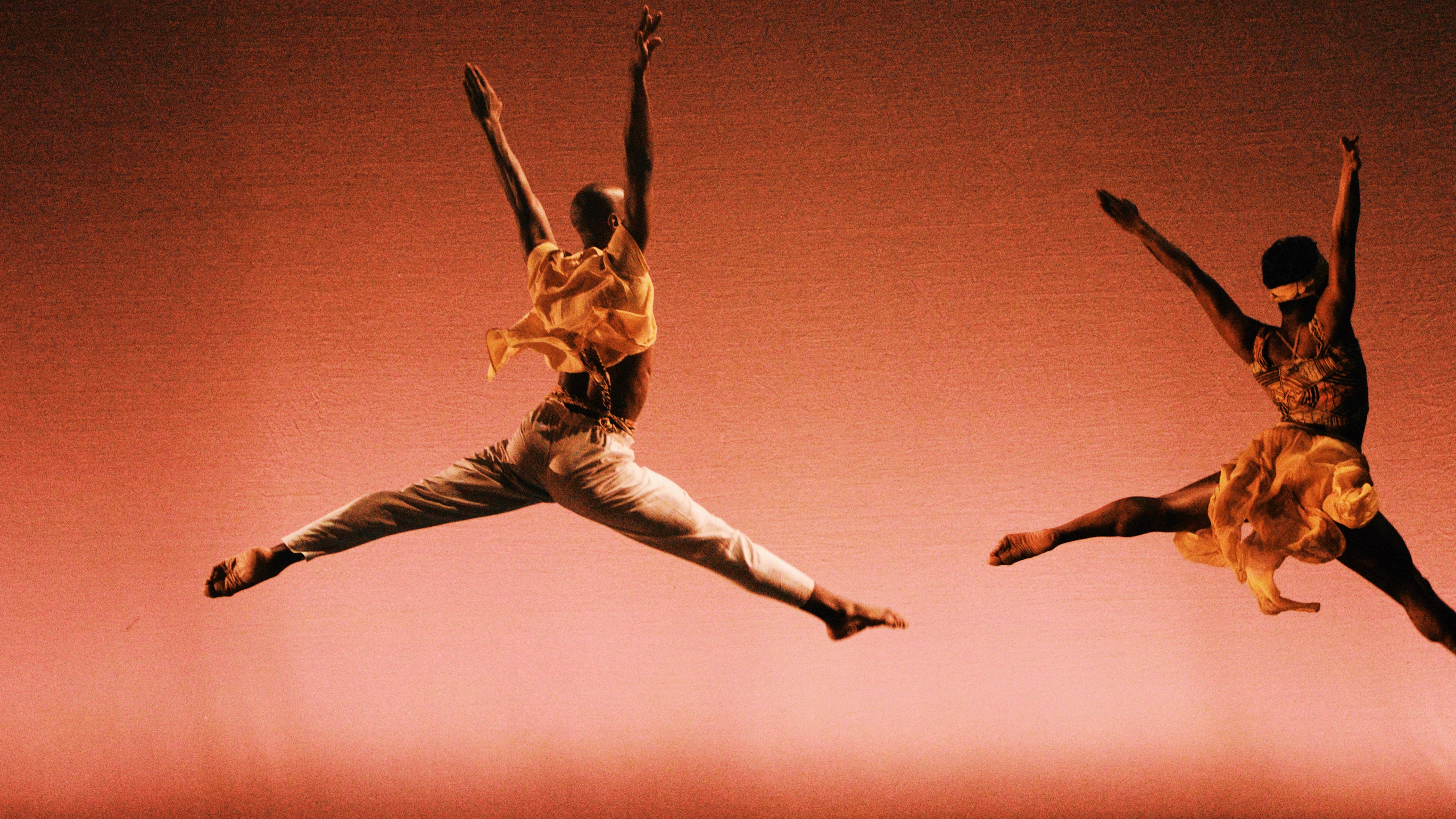 Two dancers mid-performance, one in a split leap and the other in a leap with arms raised, against a pink background.