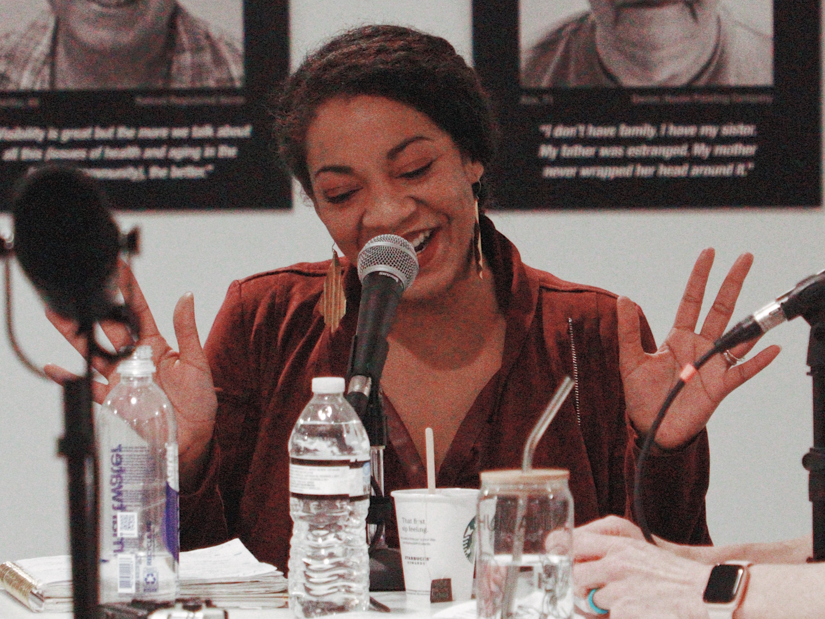 Woman speaking into microphone, smiling, with hands raised, seated at table with water bottles and cups, background with black and white photos.