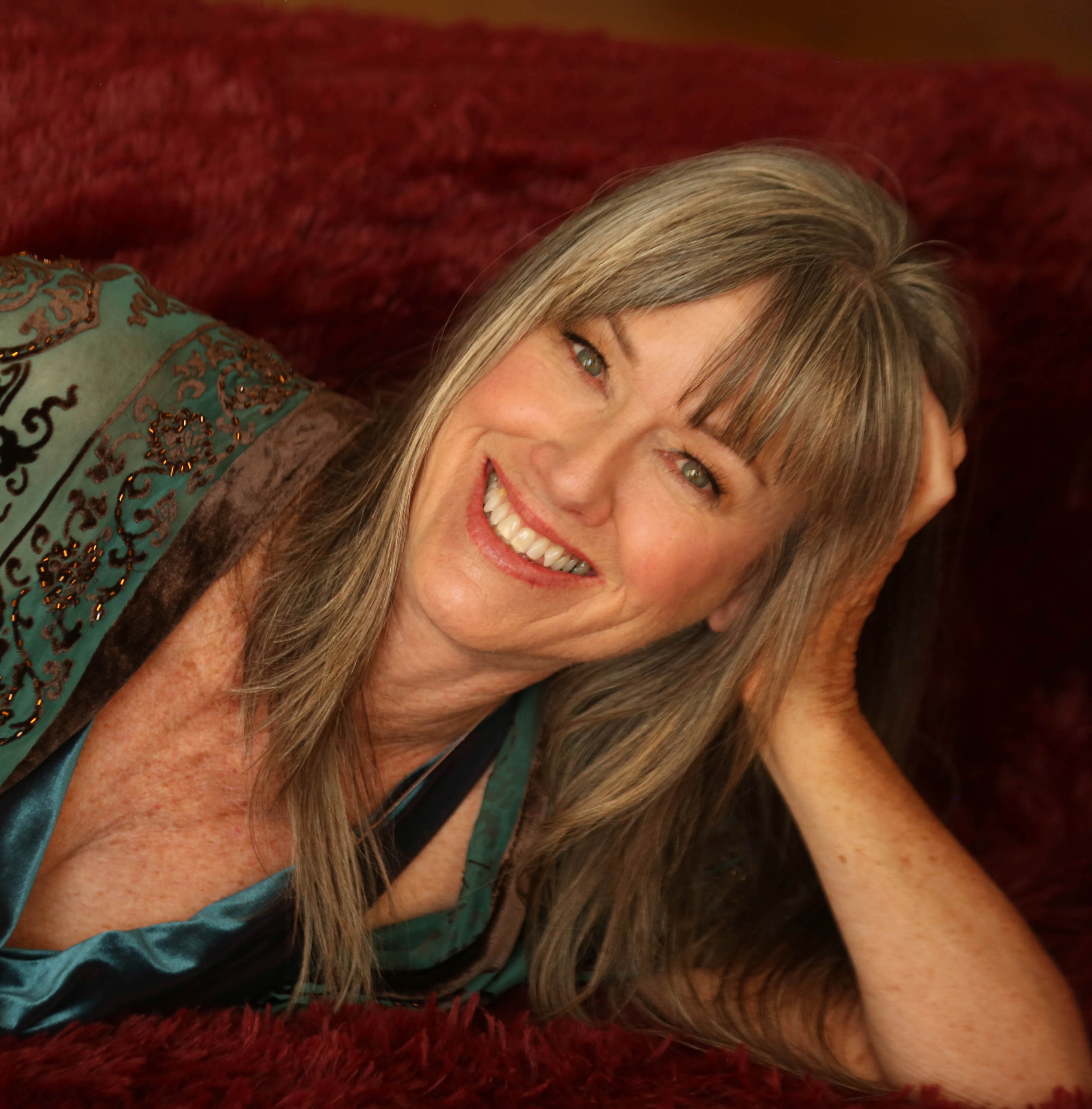 Woman with long blonde hair smiling, lying on a red surface, resting her head on her hand.