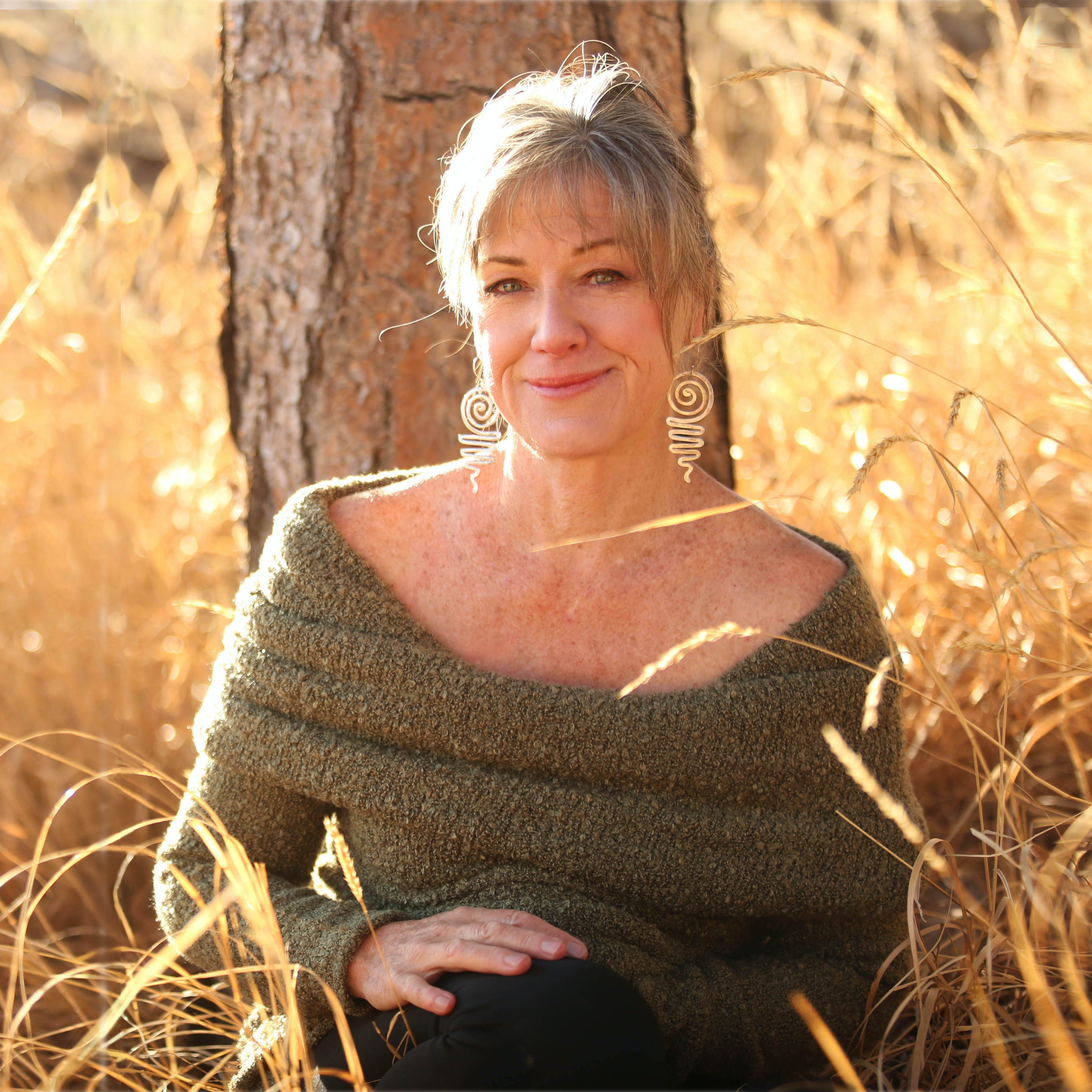 Woman with short gray hair sitting outdoors among tall golden grass, smiling, wearing a green off-the-shoulder sweater, in sunlight.
