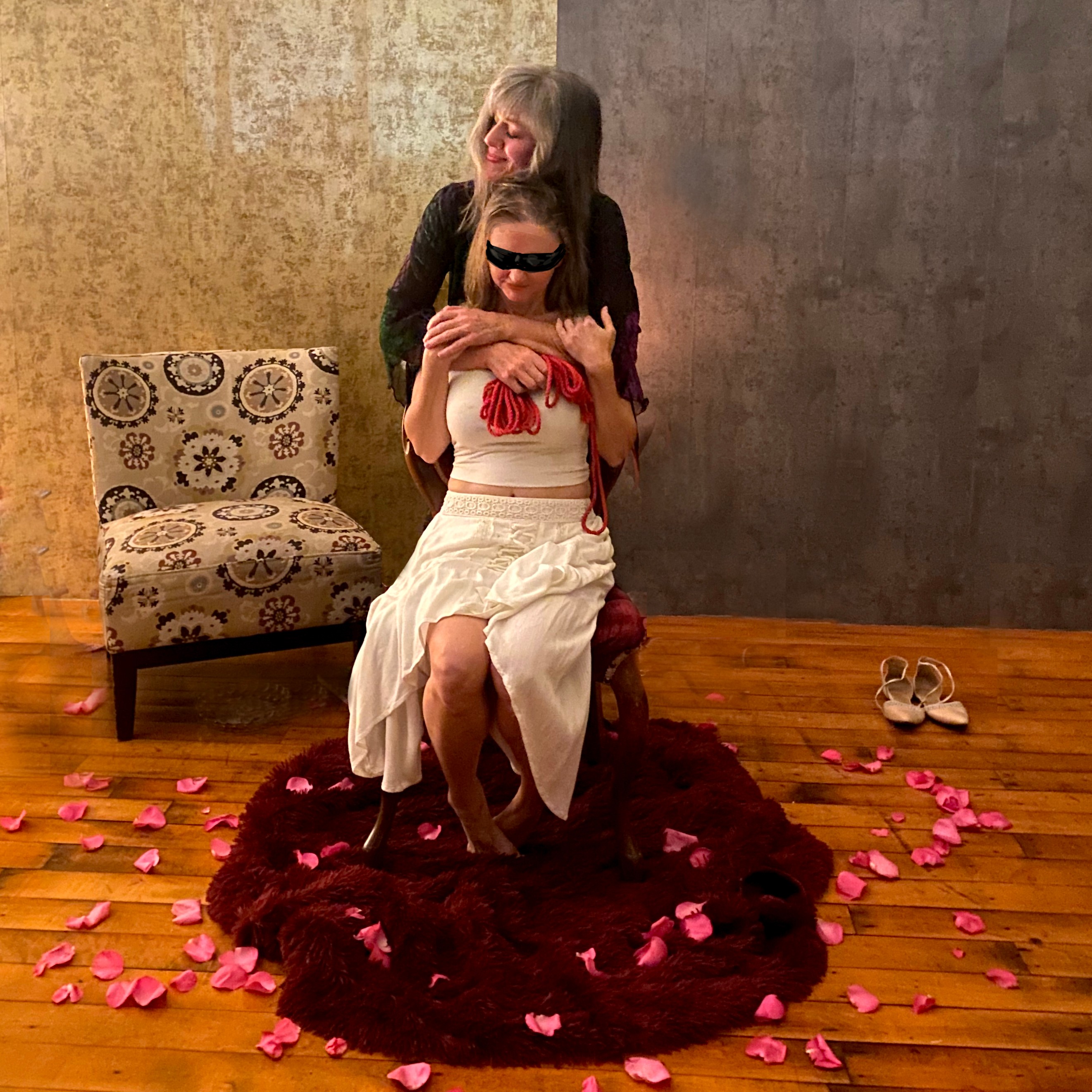 Two women, one sitting and one standing behind, in a room with a patterned chair, pink rose petals on floor and a dark rug.
