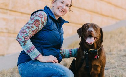 Woman kneeling beside a brown dog outdoors against a brick wall, smiling, with dry grass on the ground.
