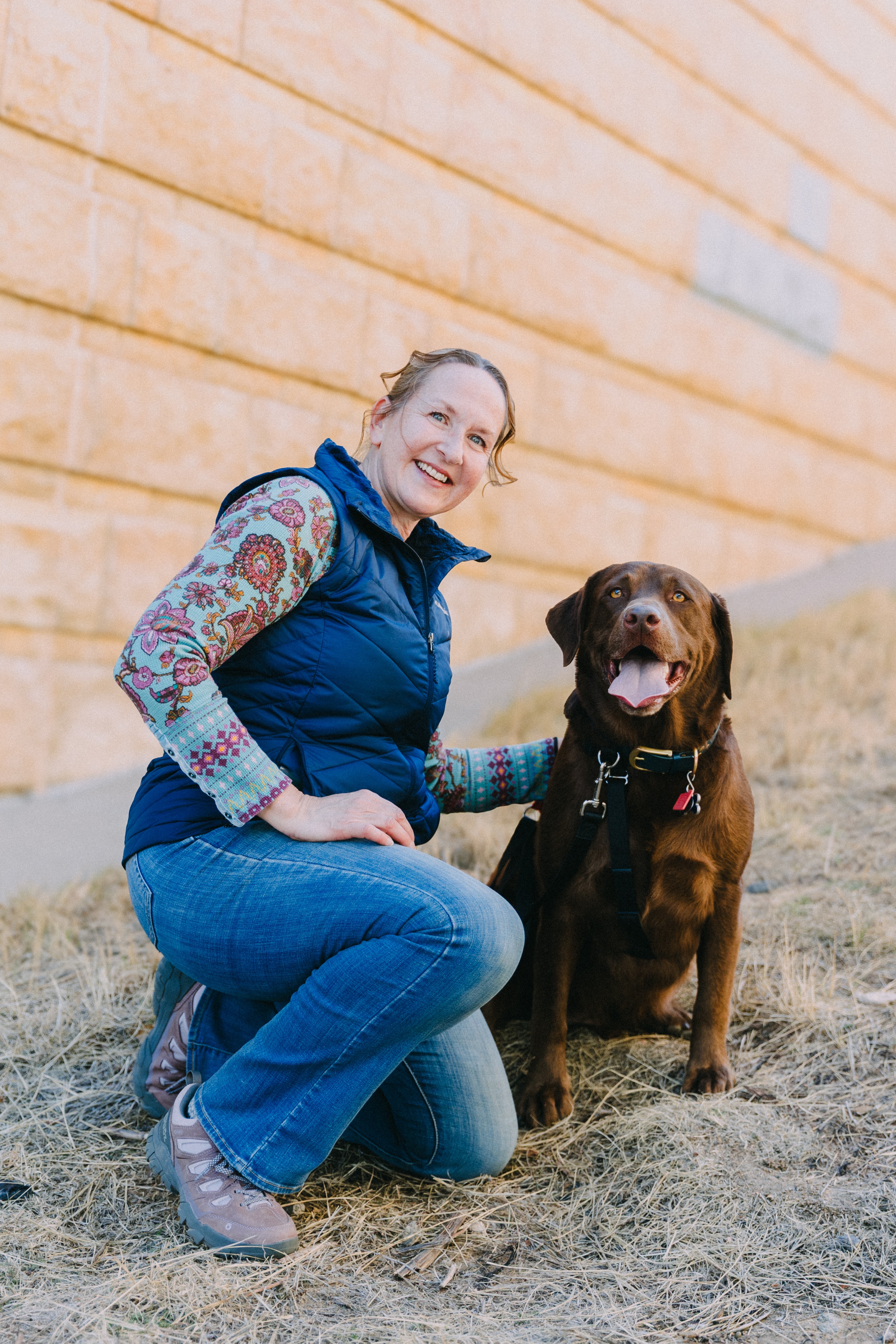 Woman kneeling beside a brown dog outdoors against a brick wall, smiling, with dry grass on the ground.