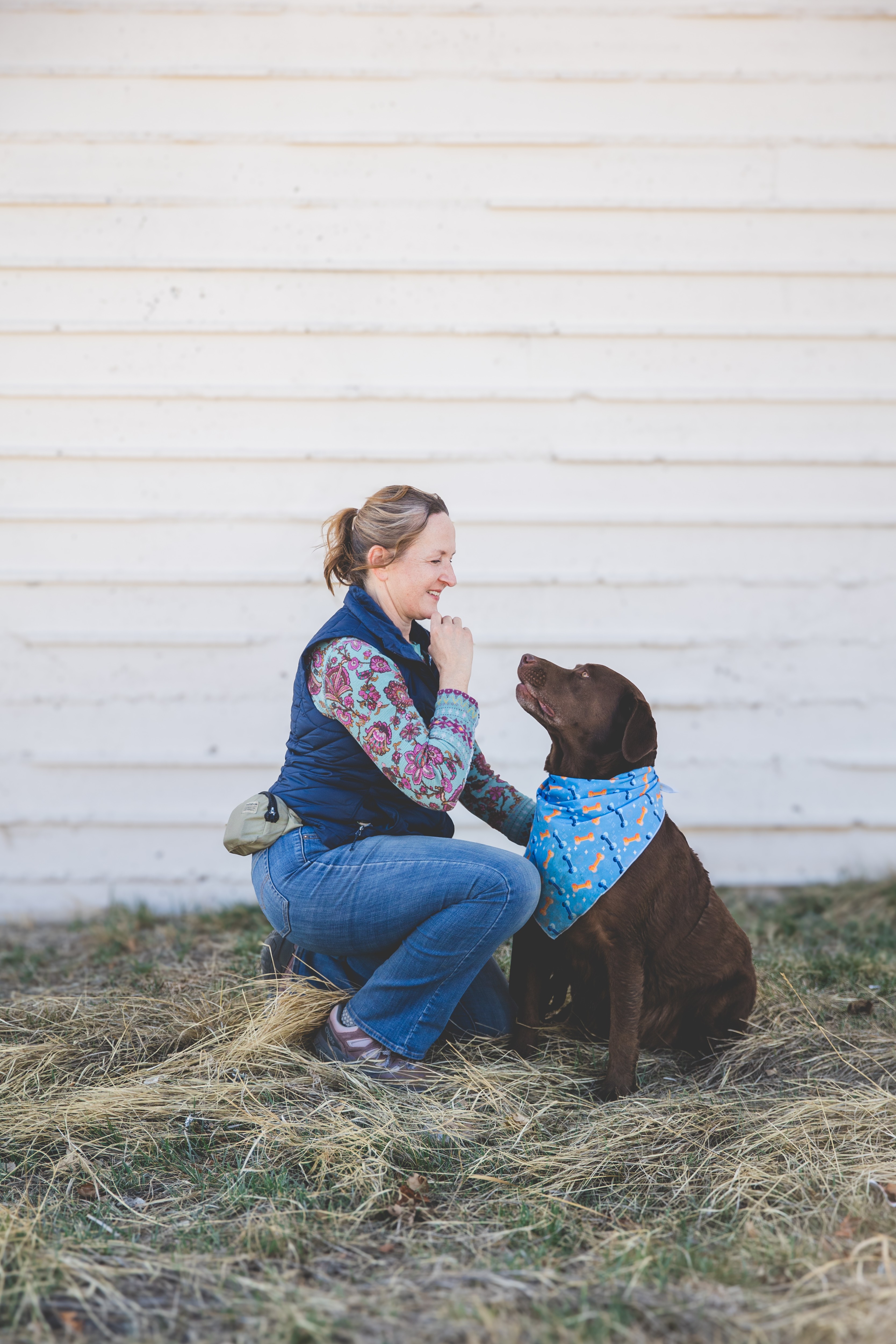 Woman kneeling and smiling at a brown dog wearing a blue bandana outdoors against a white wall.