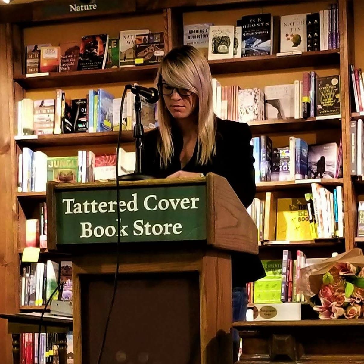 Woman with glasses at a podium in a bookstore, surrounded by shelves of books.