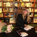Woman with glasses sitting at a table in a bookstore, surrounded by books and a bouquet of flowers.