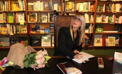 Woman with glasses sitting at a table in a bookstore, surrounded by books and a bouquet of flowers.
