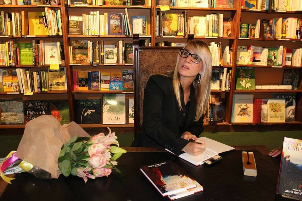 Woman with glasses sitting at a table in a bookstore, surrounded by books and a bouquet of flowers.