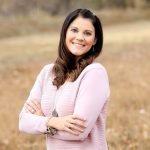 Smiling woman with long dark hair in a pink sweater standing outdoors with arms crossed, background of dry grass.