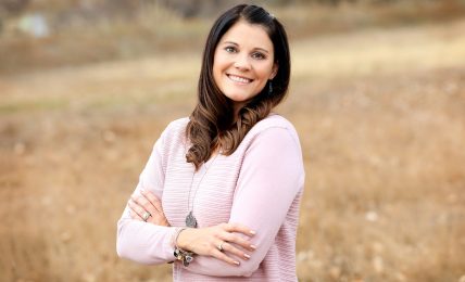 Smiling woman with long dark hair in a pink sweater standing outdoors with arms crossed, background of dry grass.