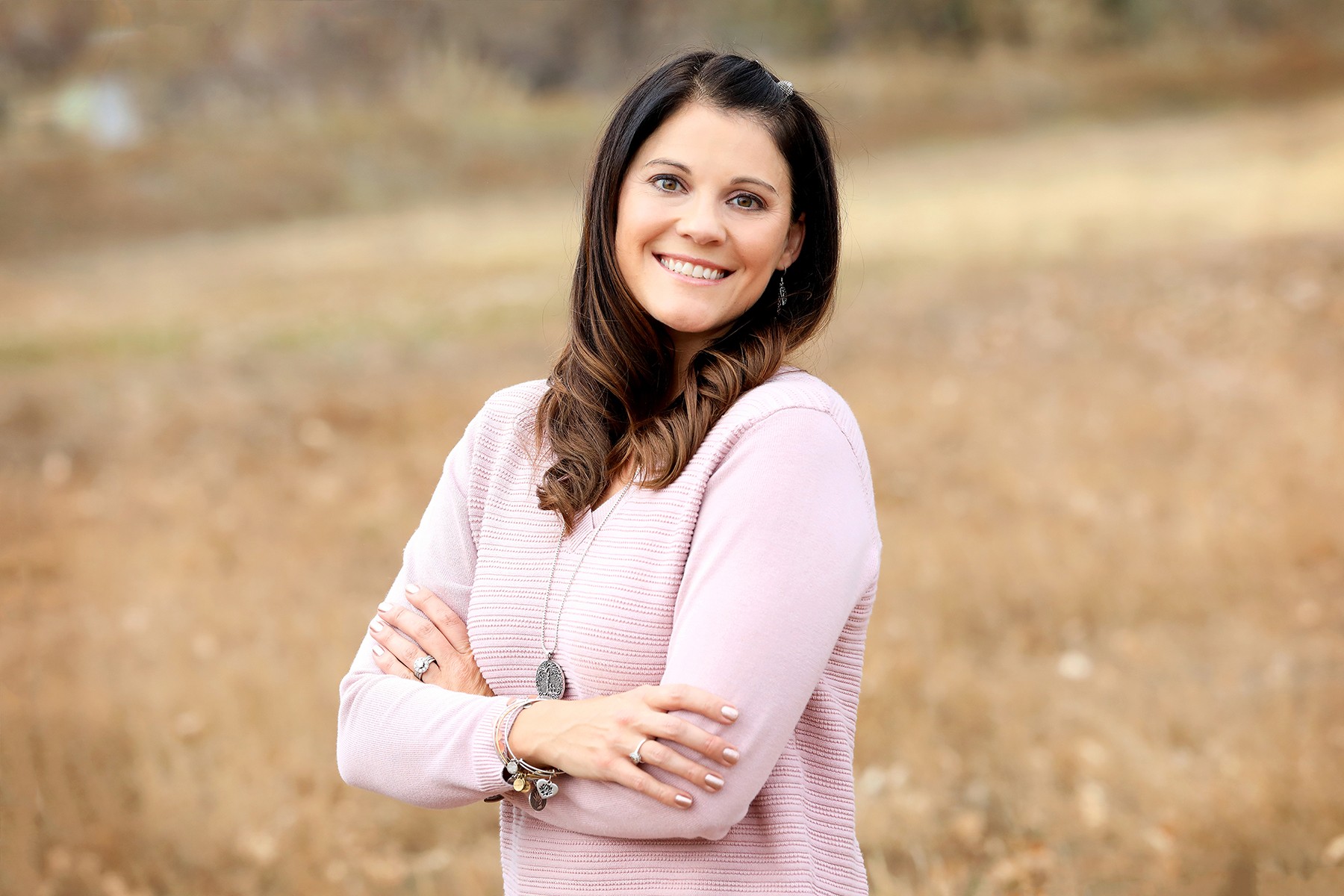 Smiling woman with long dark hair in a pink sweater standing outdoors with arms crossed, background of dry grass.