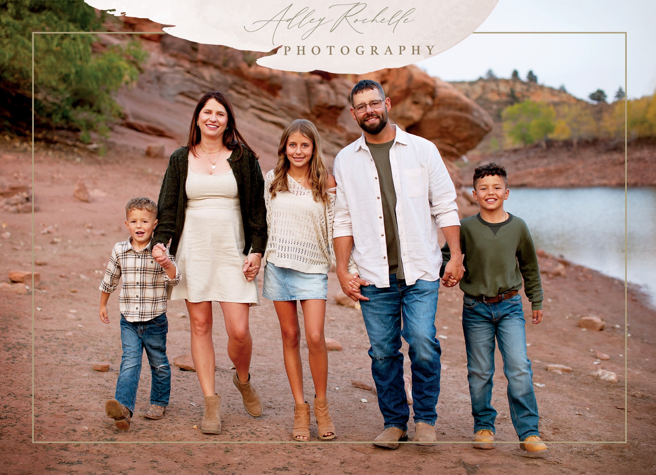 Family of five walking outdoors near water, with rocky landscape and trees in background, smiling and holding hands.