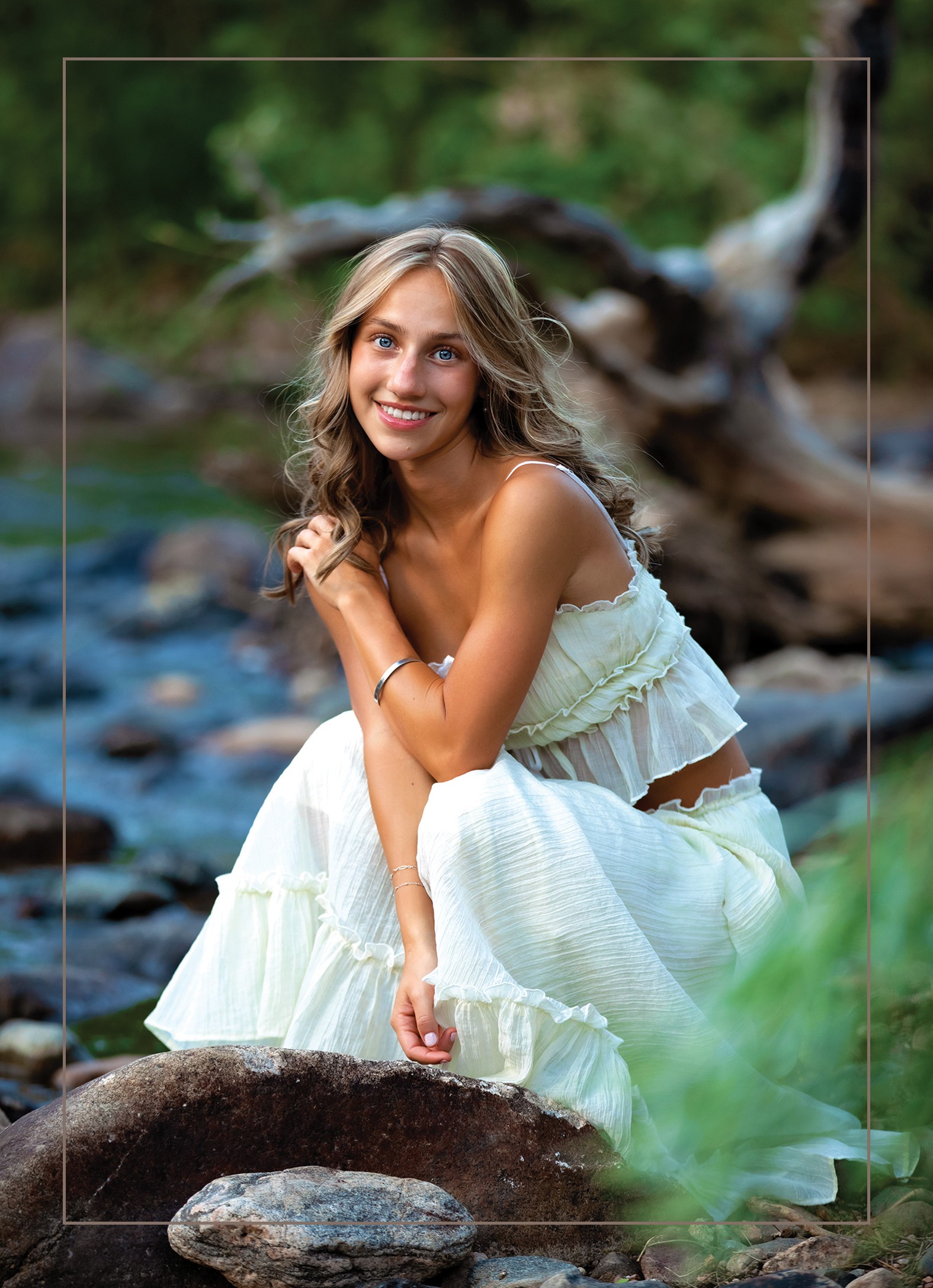 Young woman in white dress sitting on a rock near a river, smiling, with trees and driftwood in background.