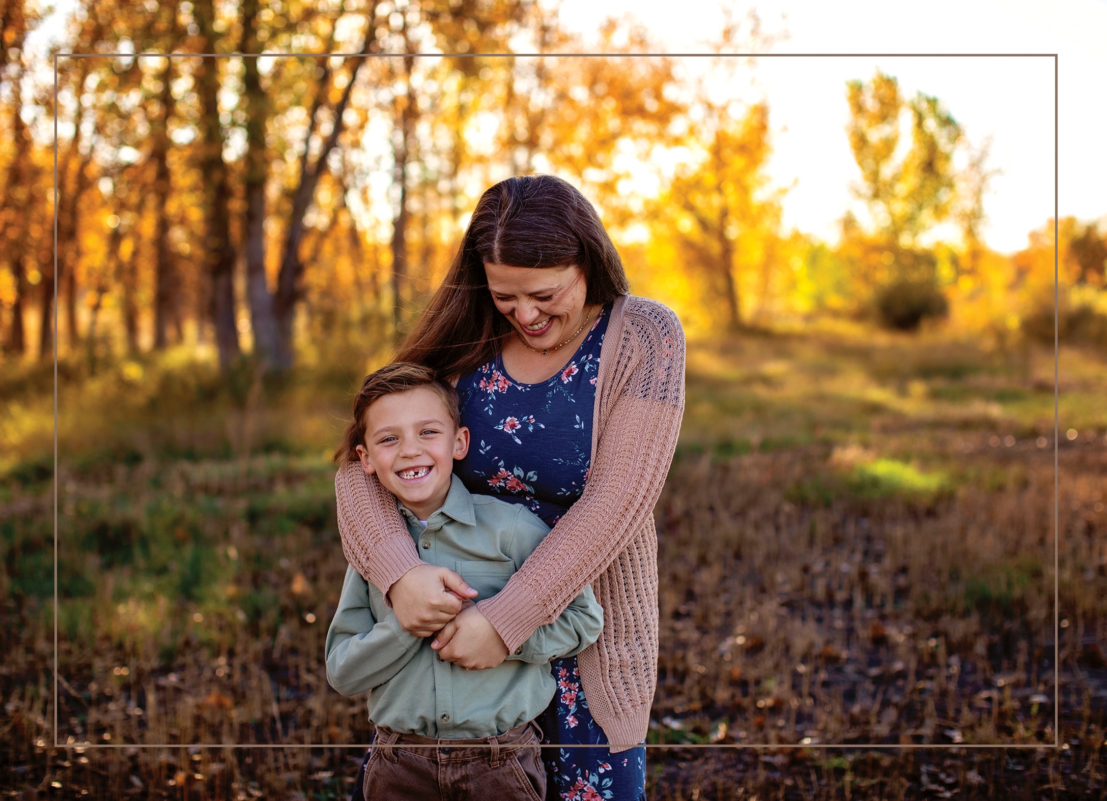 Woman hugging a smiling boy outdoors during autumn, trees with orange leaves in background.