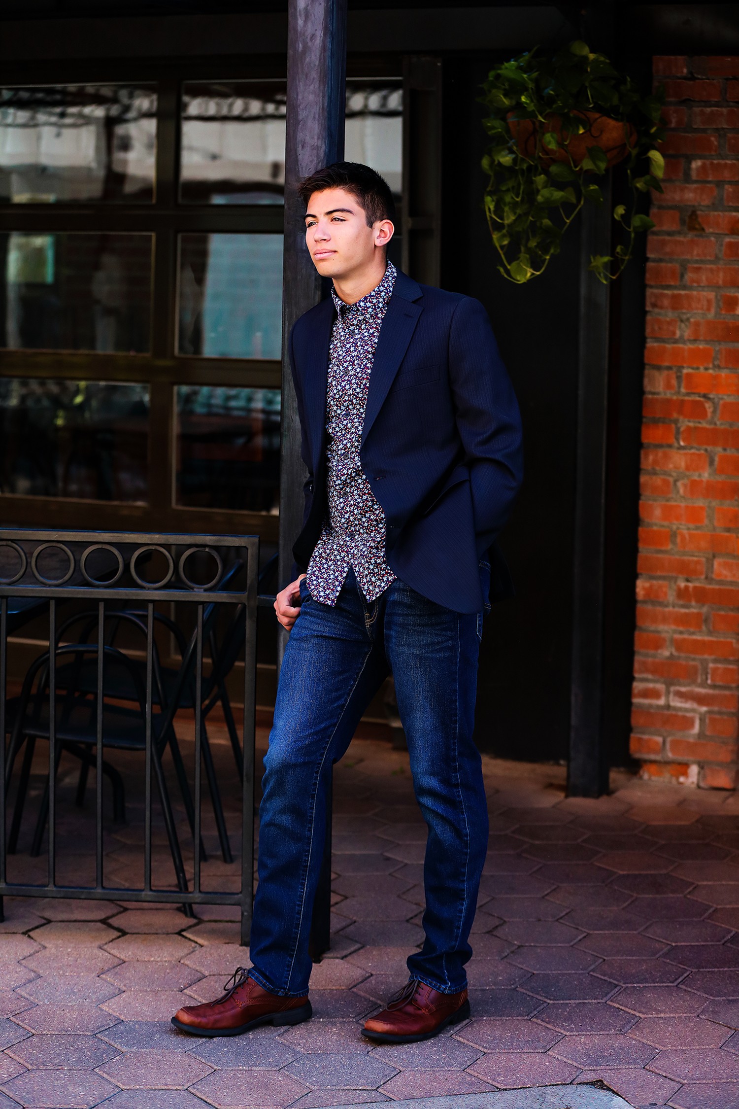 Young man in a navy blazer and patterned shirt standing outside near a brick wall and window.