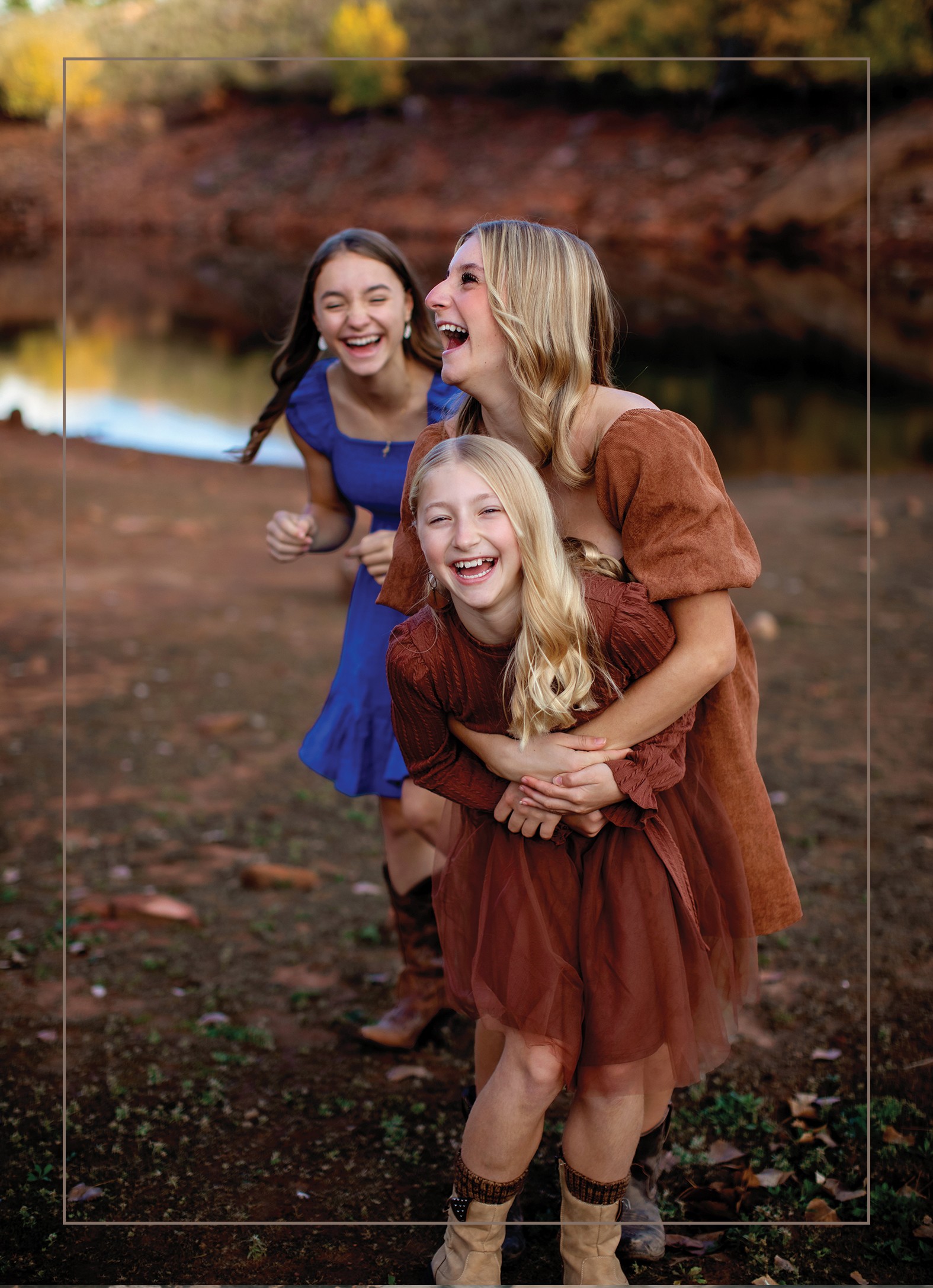 Three girls laughing outdoors near a body of water, with trees and a rocky bank in the background.